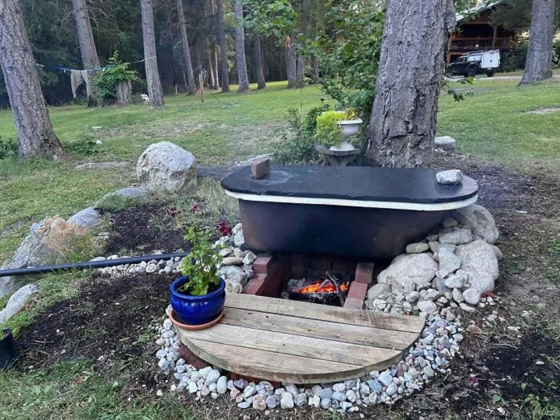 Bathtub on bricks over fire pit with a wooden cover and potted plant nearby.