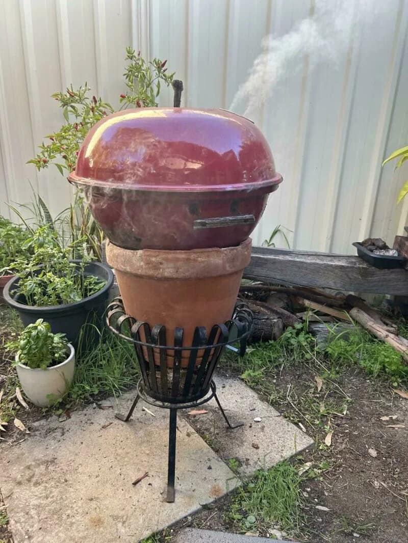 Barbecue grill on a stand with a flower pot underneath, smoke rising from it.