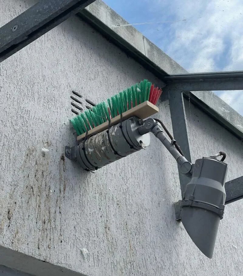 Unusual device with a broom attached to a pipe on a building wall.