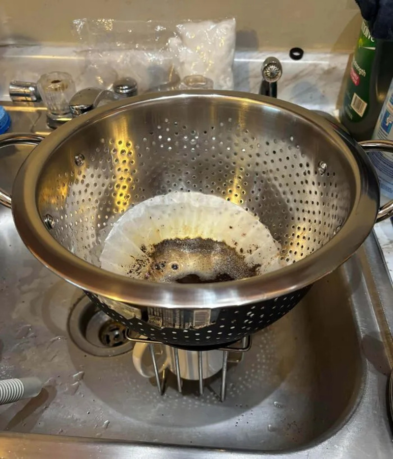 A colander in a sink with burnt coffee grounds and a paper filter inside.