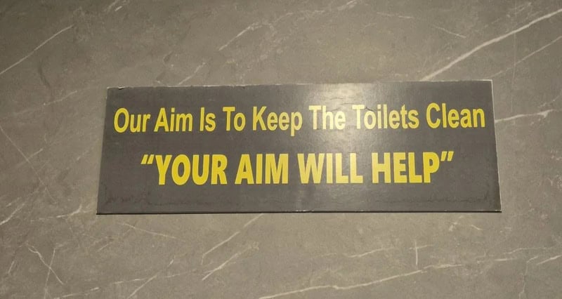 Sign reading 'Our Aim Is To Keep The Toilets Clean "YOUR AIM WILL HELP"' on a gray tiled floor.