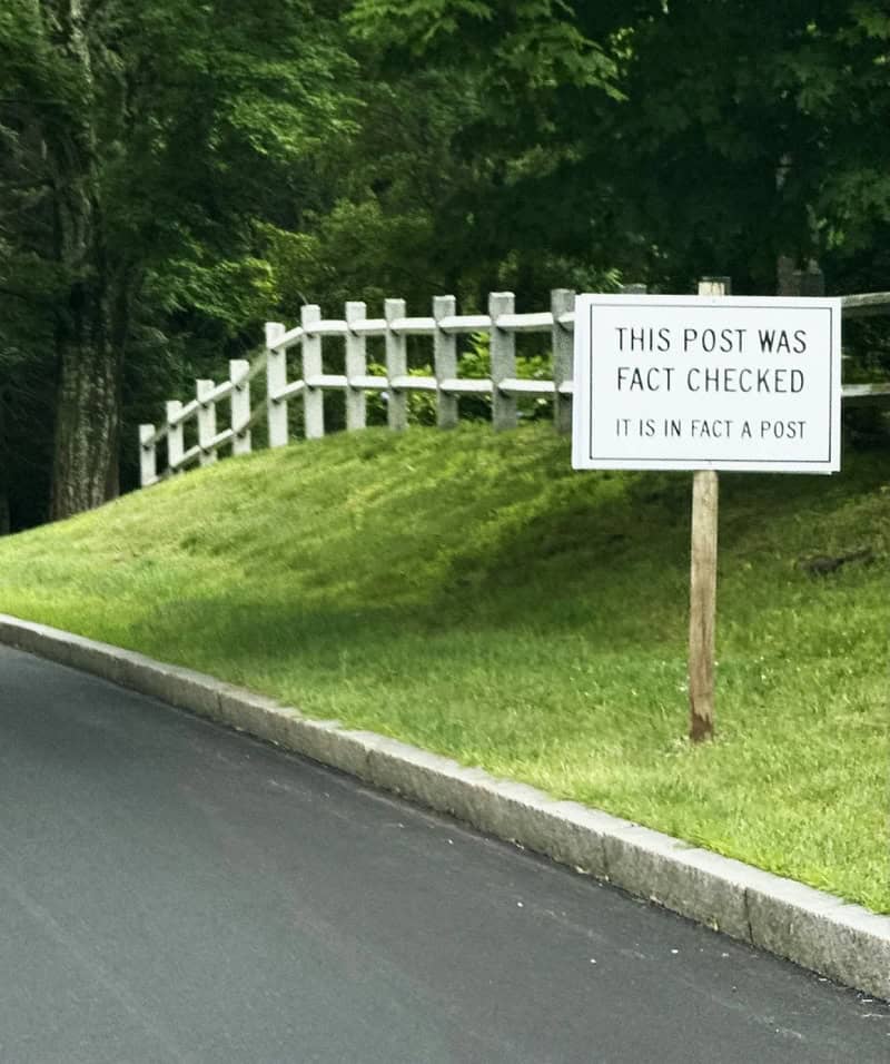 Sign reads 'This post was fact checked. It is in fact a post.' near a white fence and green grass.