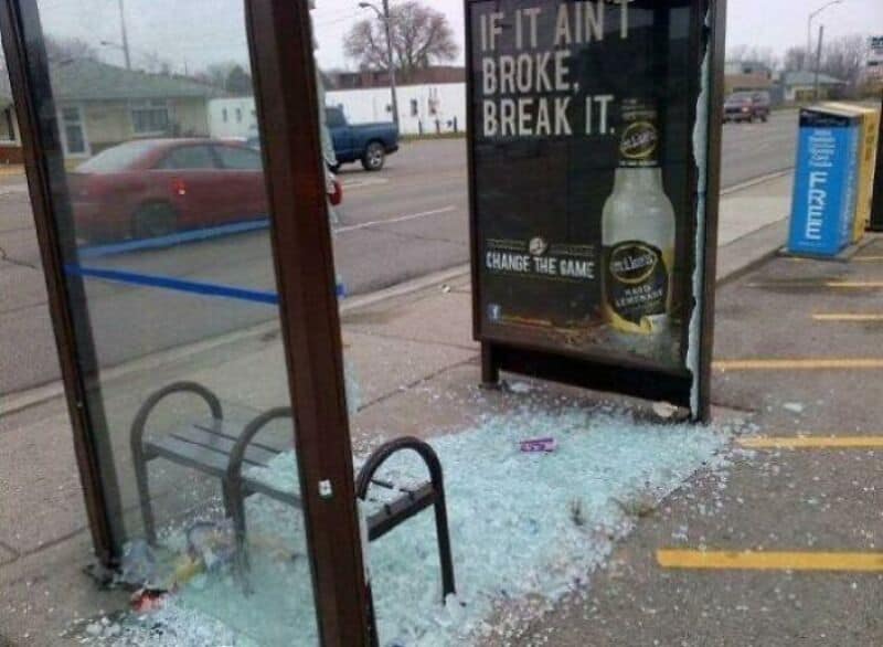 Broken glass at bus stop with a beer ad and a blue newspaper box nearby.