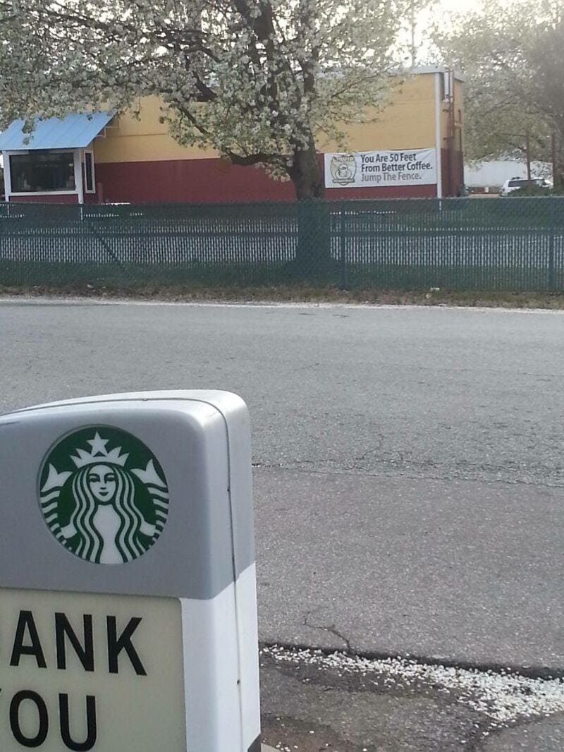 Sign with Starbucks logo and partial message, billboard in background with tree and fence.