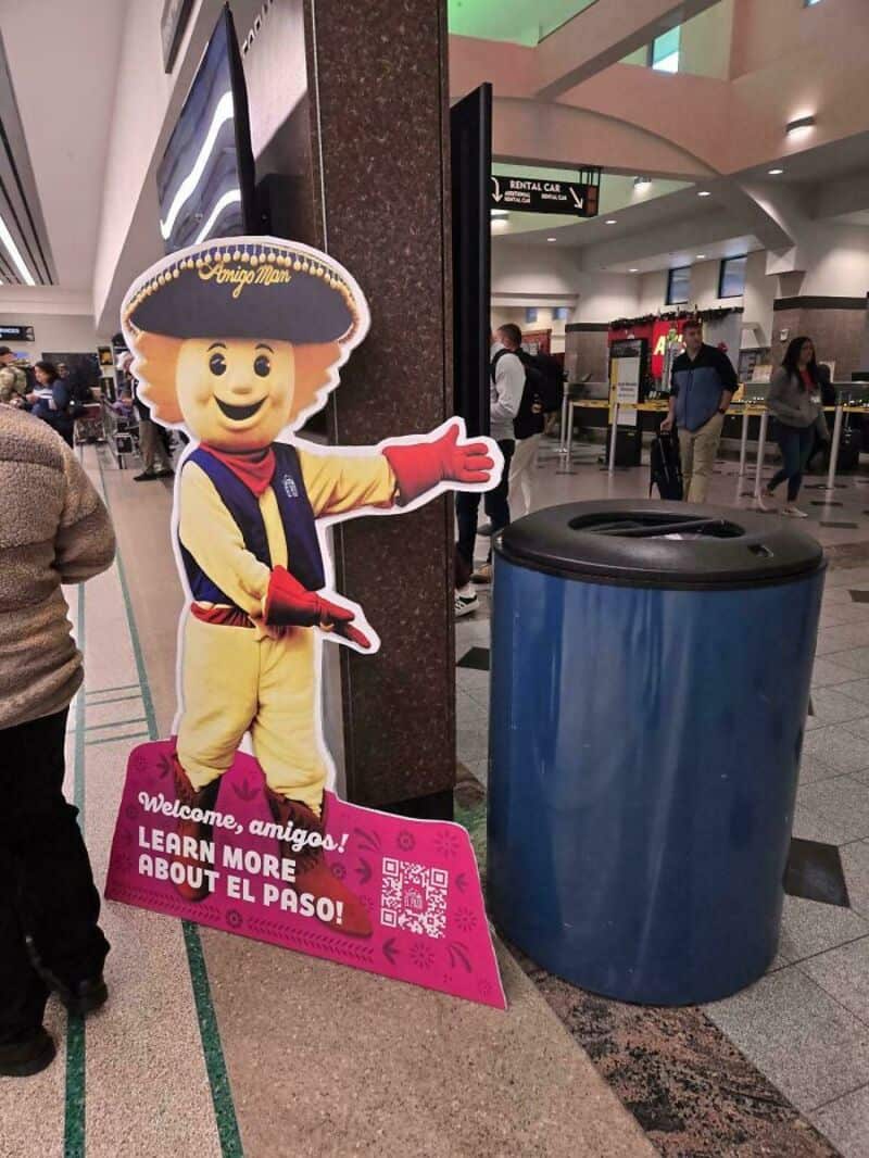 Costumed character at airport with sign about El Paso in Spanish and English.