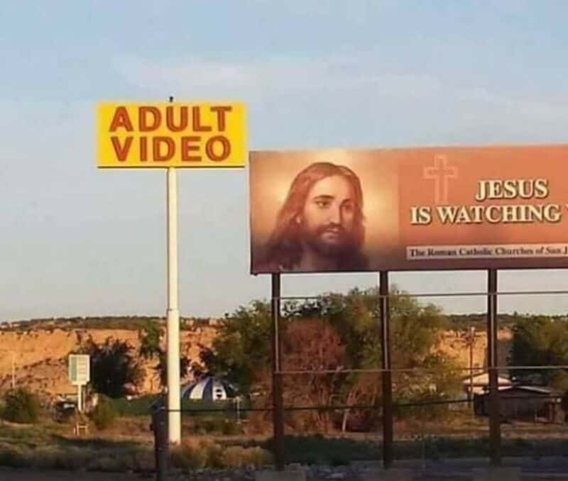 Billboard with Jesus image and text, 'JESUS IS WATCHING', and a yellow sign 'ADULT VIDEO'.