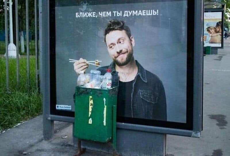 Man with a beard holding chopsticks near a trash bin with water bottles.