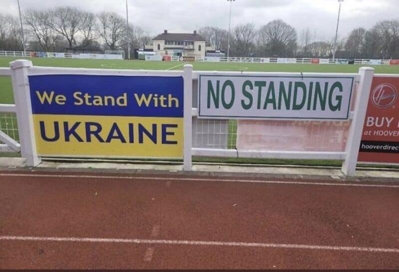 We Stand With UKRAINE NO STANDING sign on a fence at a sports field.