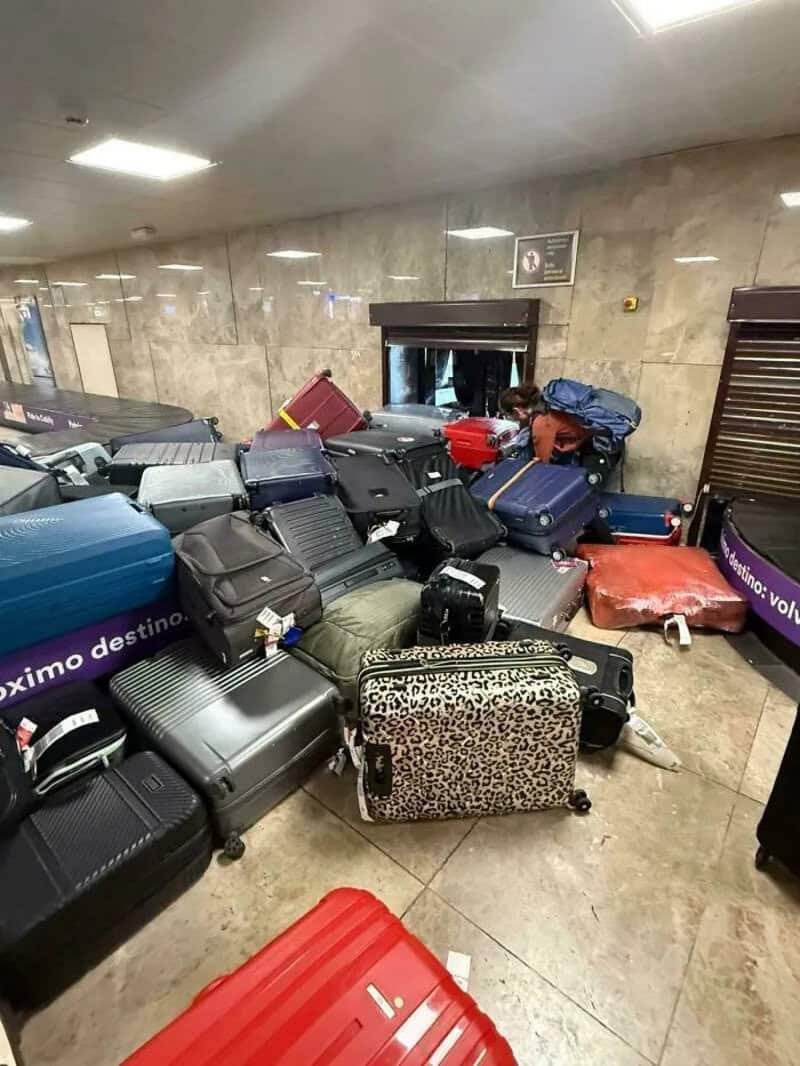 Luggage scattered in an airport baggage claim area, some falling off the conveyor.