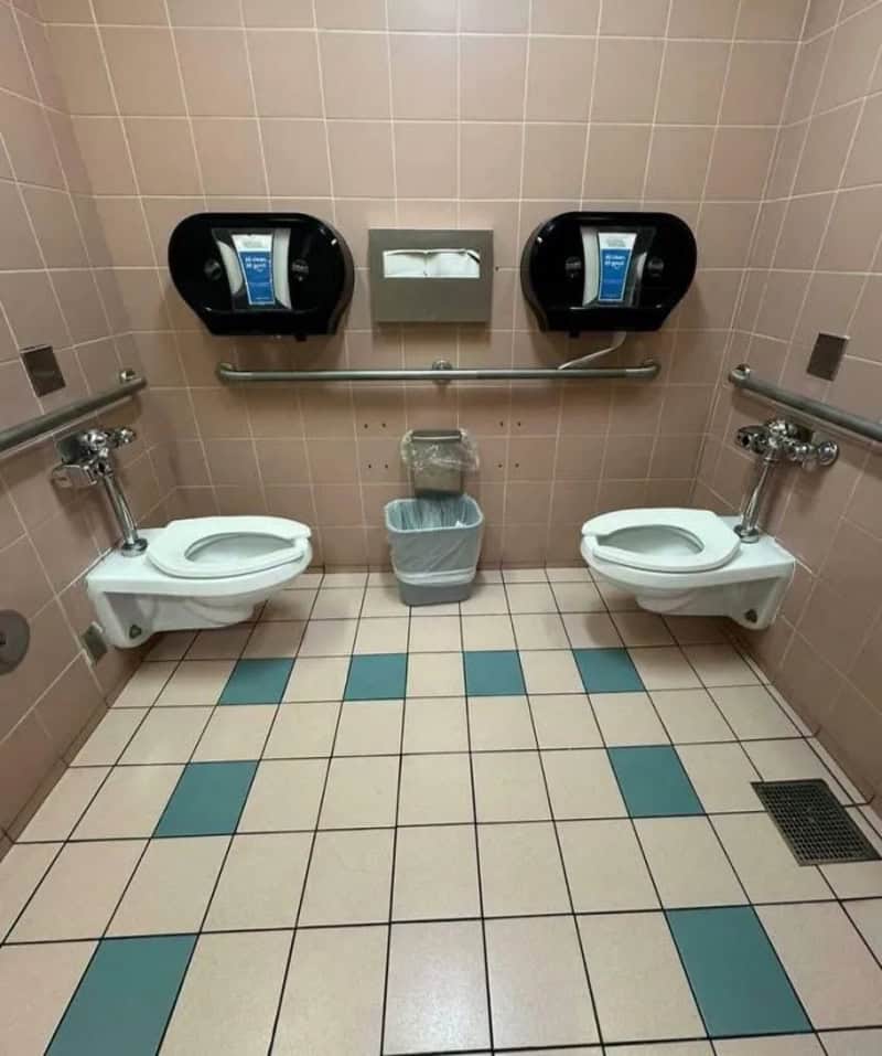 Two hand dryers above two urinals in a bathroom with a trash bin in the middle.