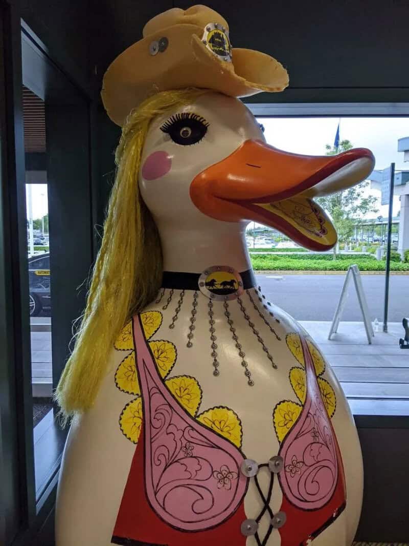 Large duck sculpture with a hat, long blonde hair, and a pink bra, inside a building.
