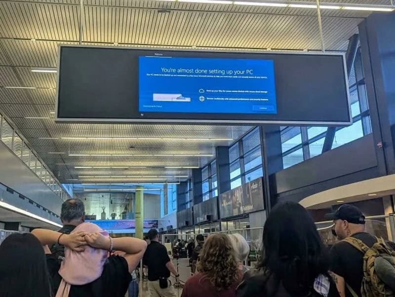 People waiting at an airport with a large screen showing PC setup message.