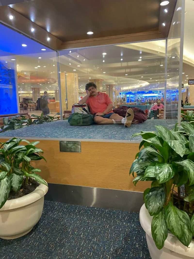 Man lying on a glass platform at airport, surrounded by plants, with travelers in background.