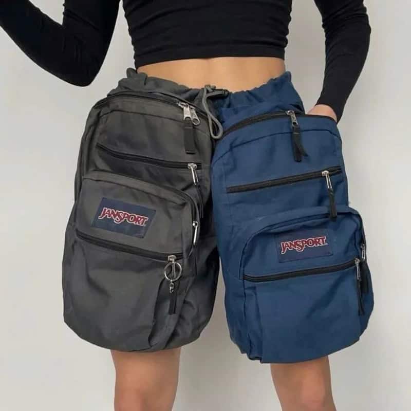 Two women wear mismatched JanSport backpacks, one gray and one blue, with multiple zippers.