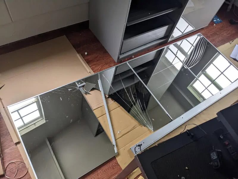 Broken mirror on a wooden floor with furniture and a window reflection.