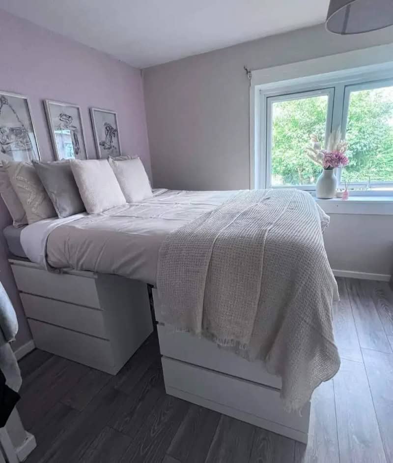 A bed with storage drawers under it in a bedroom with a window and pink wall art.