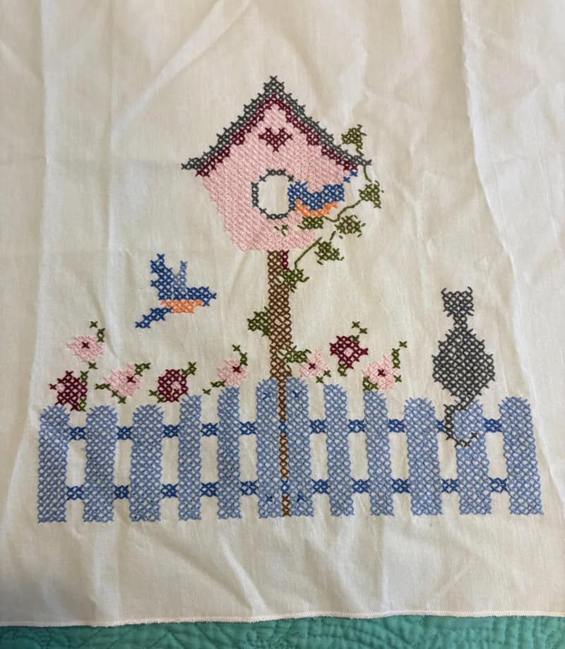 Cross-stitch embroidery of a birdhouse, birds, and a fence on fabric.
