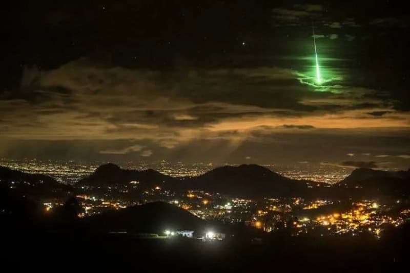 Green meteor streaks across night sky over city and mountains.
