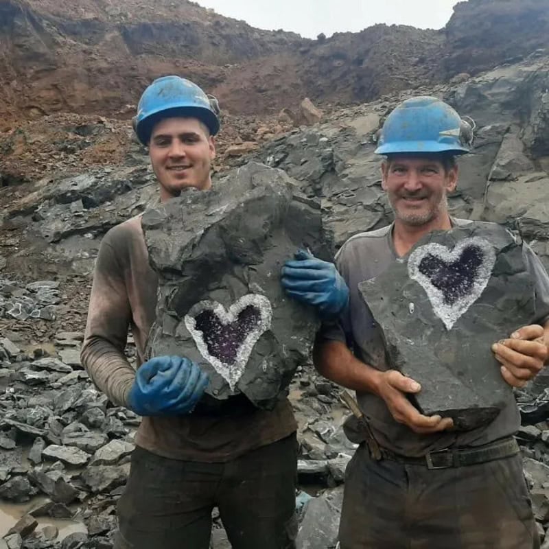 Two miners in helmets holding rocks with heart-shaped designs in a quarry.