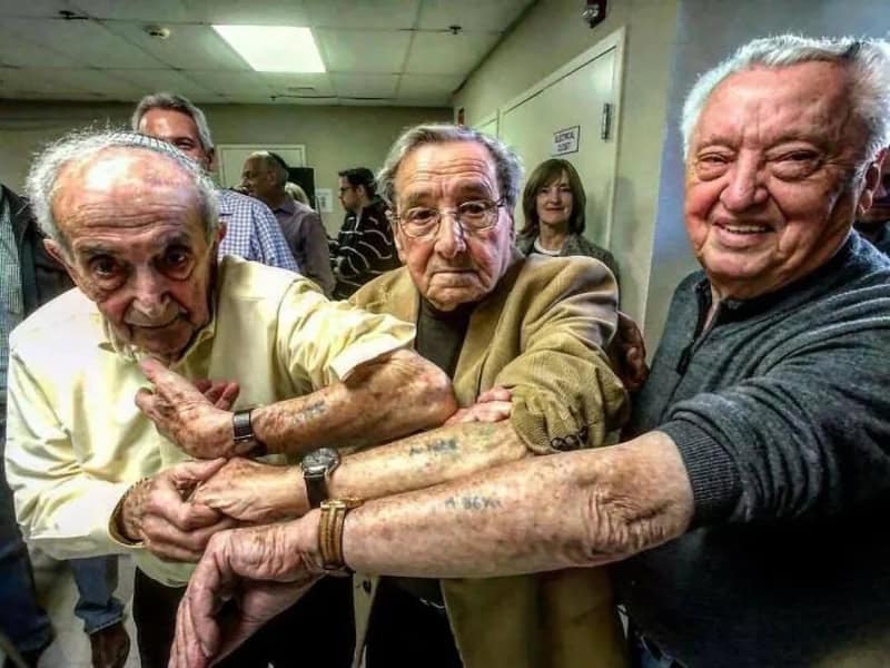 Three elderly men holding hands together in a room with other people in the background.