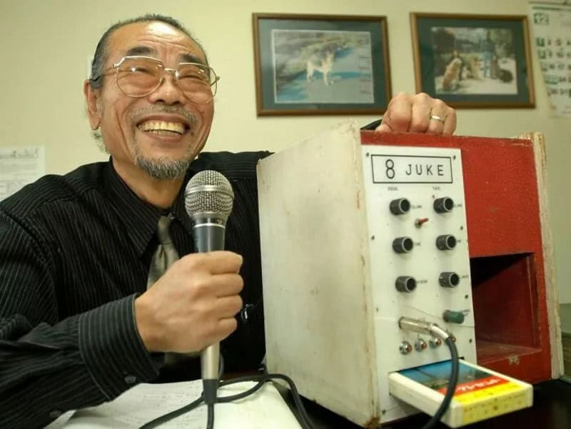 Smiling man with glasses holding a microphone next to a vintage jukebox labeled '8 JUKE'.