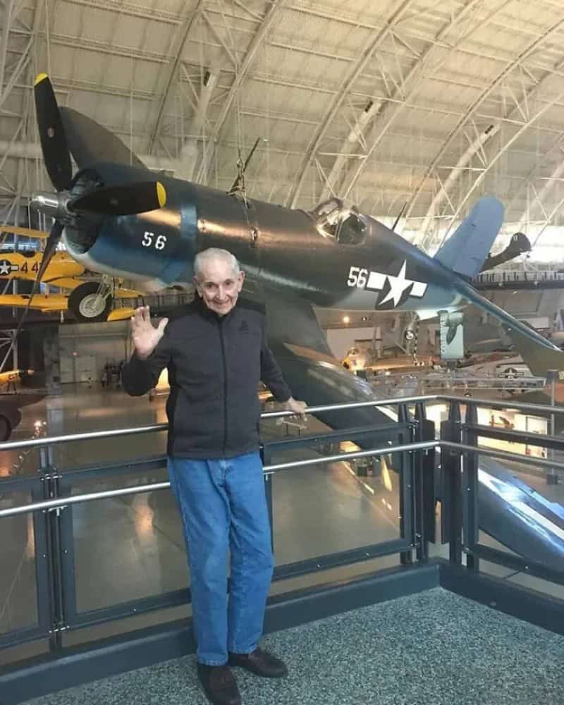 Elderly man waving in front of a vintage aircraft at an aviation museum.