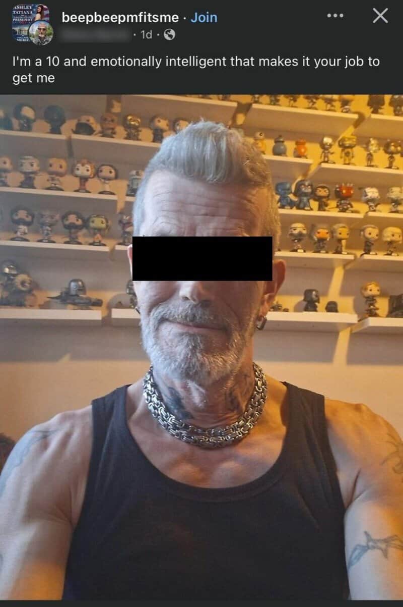 Older man with gray hair and beard, wearing a black tank top and silver chains, in front of shelves.