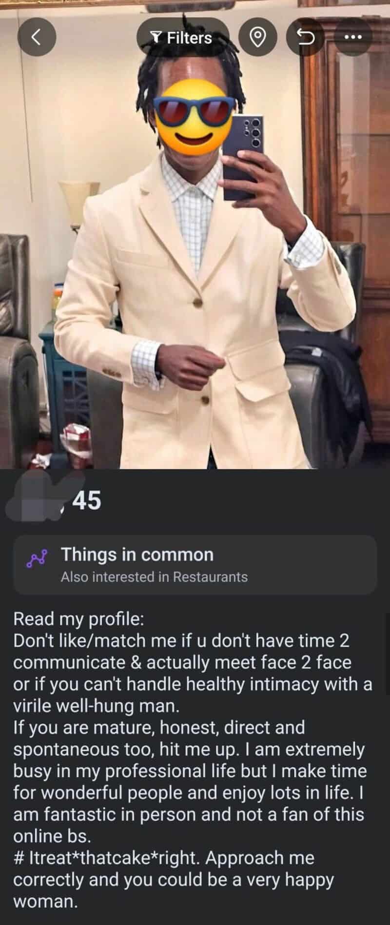 Man in beige suit with sunglasses emoji on face, taking mirror selfie.