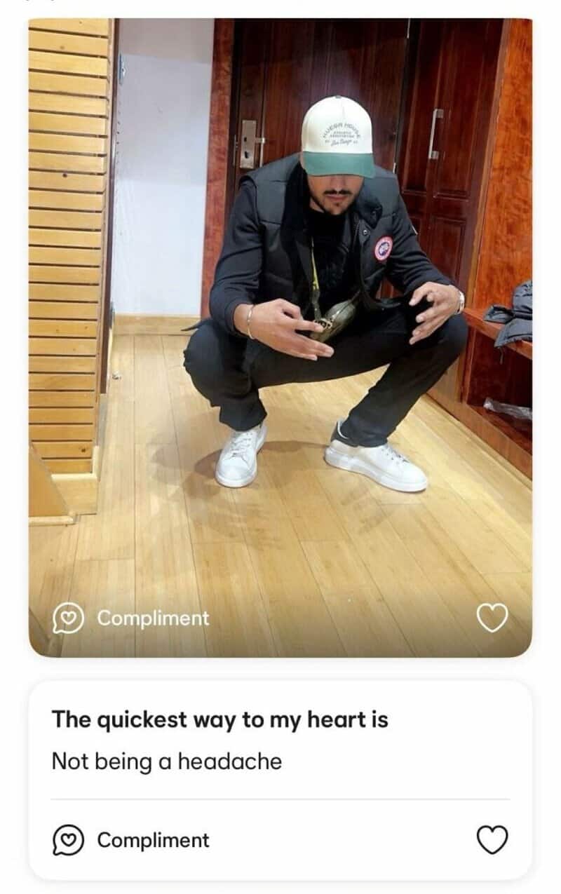 Man squatting in a room, wearing a black jacket, white cap, and white sneakers.