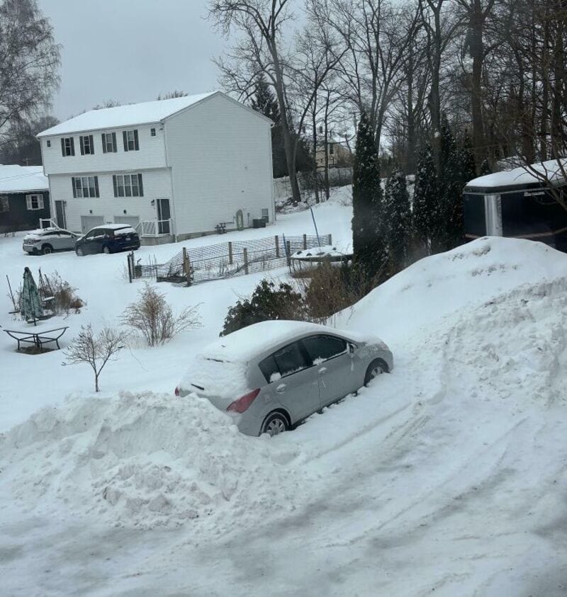 Car buried in snow next to a large snowbank, with a house and trees in the background.