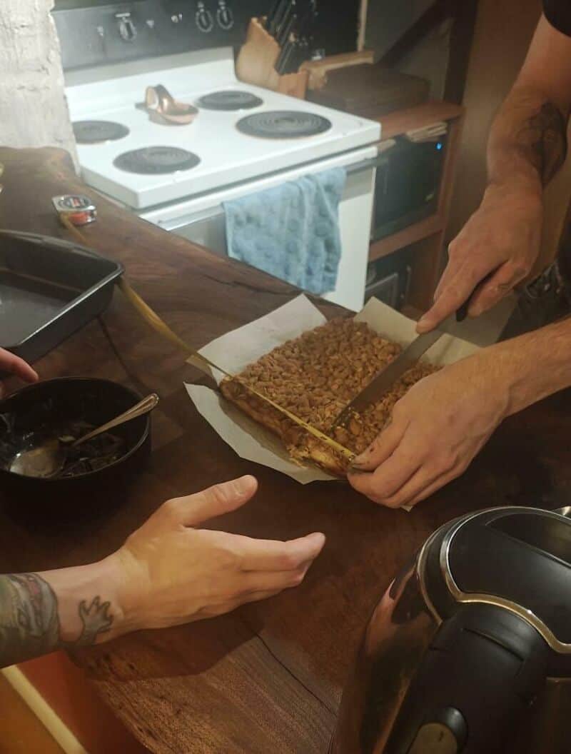 Person cutting a layered dessert with a knife and spatula in a kitchen.