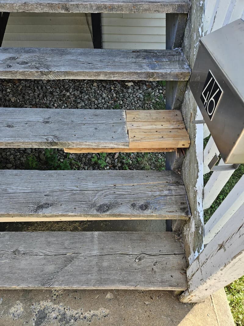 Damaged wooden stairs with one step replaced by a new piece of wood.
