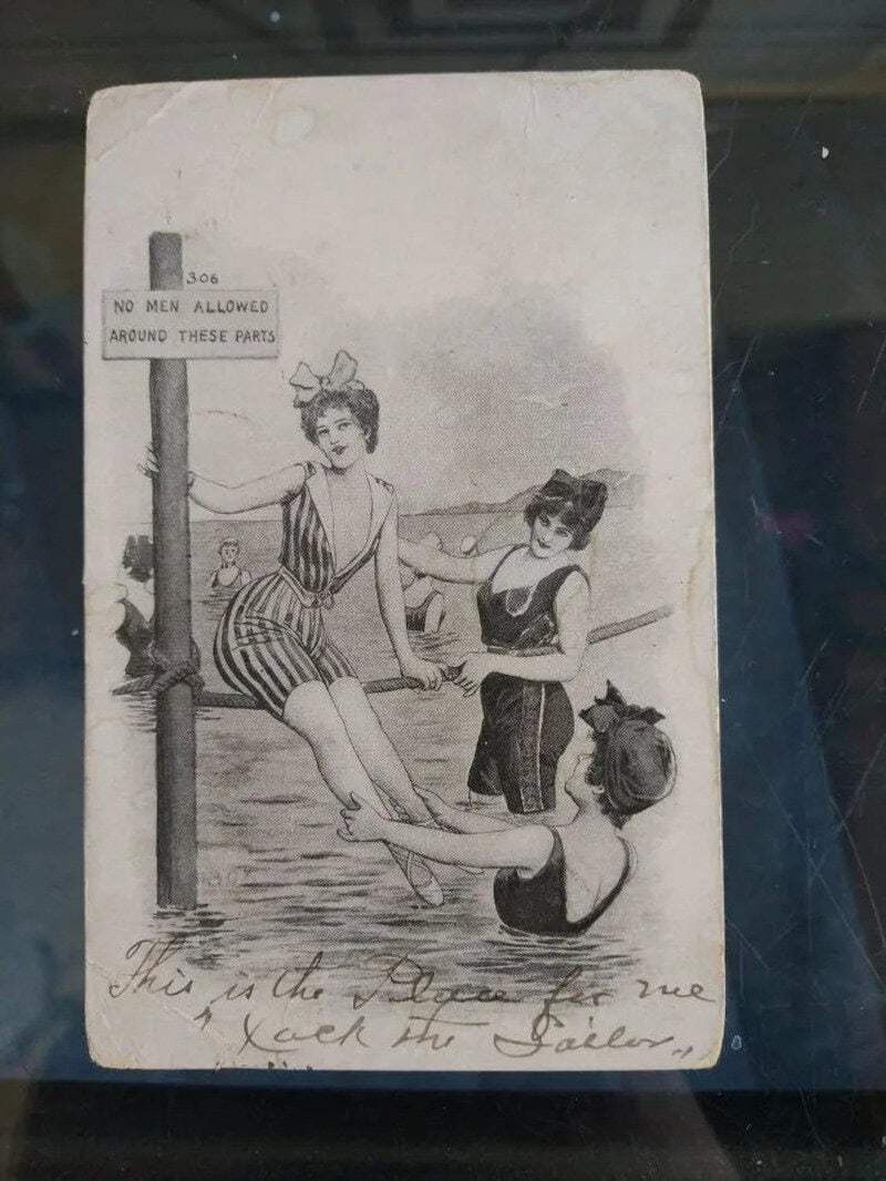Vintage black and white photo of three women in swimsuits by the water, one sitting on a pole.