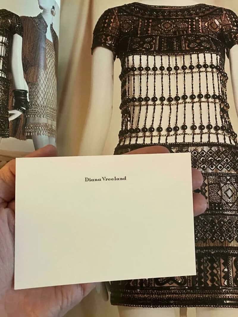 Mannequin wearing a black lace dress with beaded details, holding a white card with 'Diana Vreeland'.