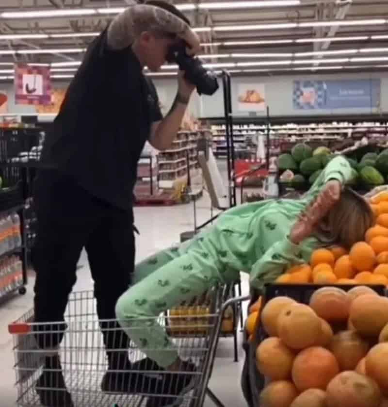 Person taking photo of woman lying on shopping cart in produce aisle.