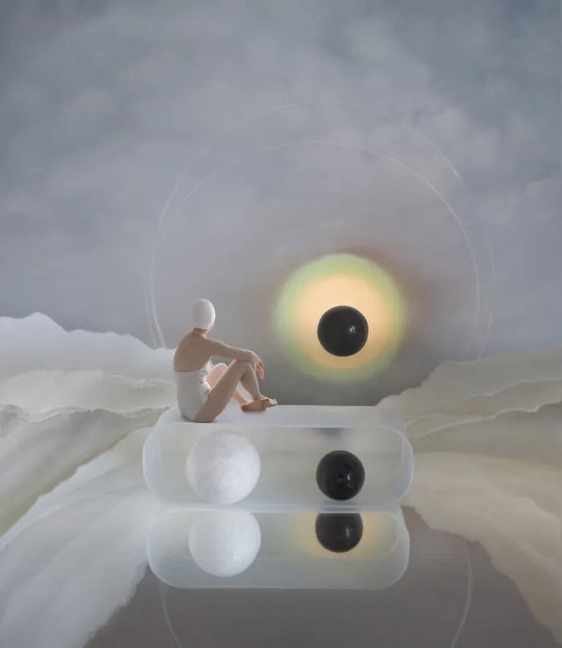 Person sitting on a white sphere with a black sphere in front, surreal landscape with a large black/.