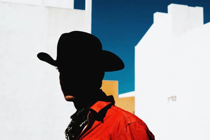 Silhouette of a person wearing a hat and red jacket against a white building and blue sky.