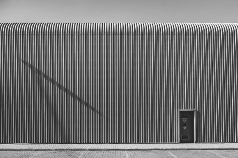 Black and white photo of a corrugated metal wall with a door and shadow.