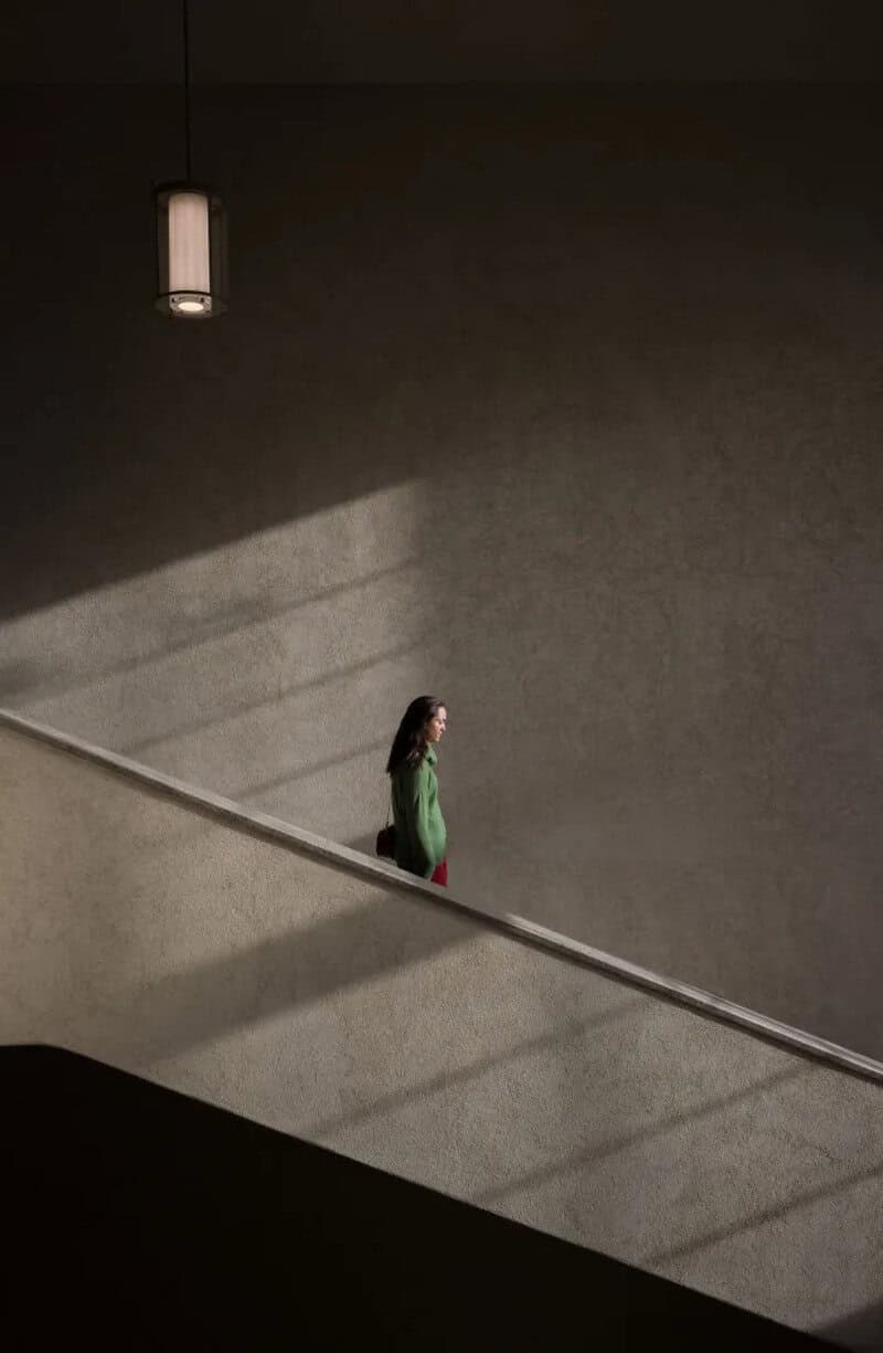 Woman in green top walking down a staircase with shadows and a lamp overhead.
