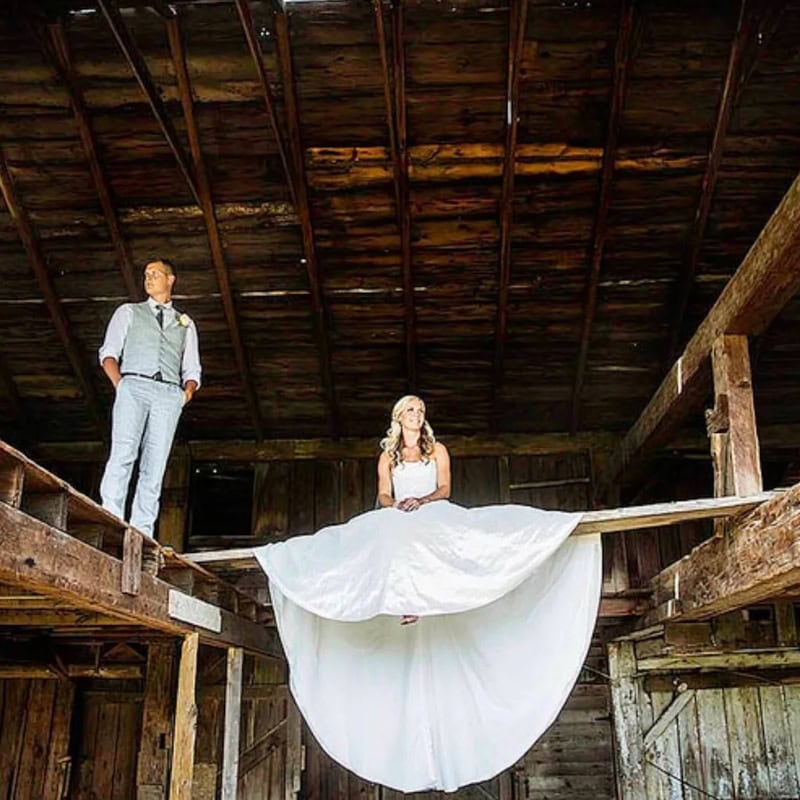 Bride sitting on a wooden beam with groom standing nearby in a rustic barn.