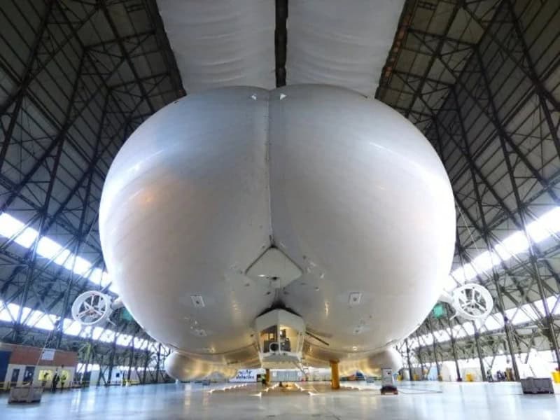 Large white airship inside a hangar with a high ceiling and metal framework.