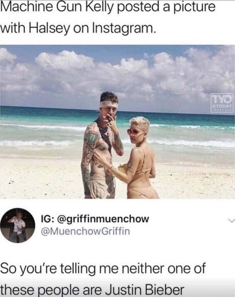 Two people on a beach, one with tattoos and sunglasses, the other with short blonde hair, holding a.