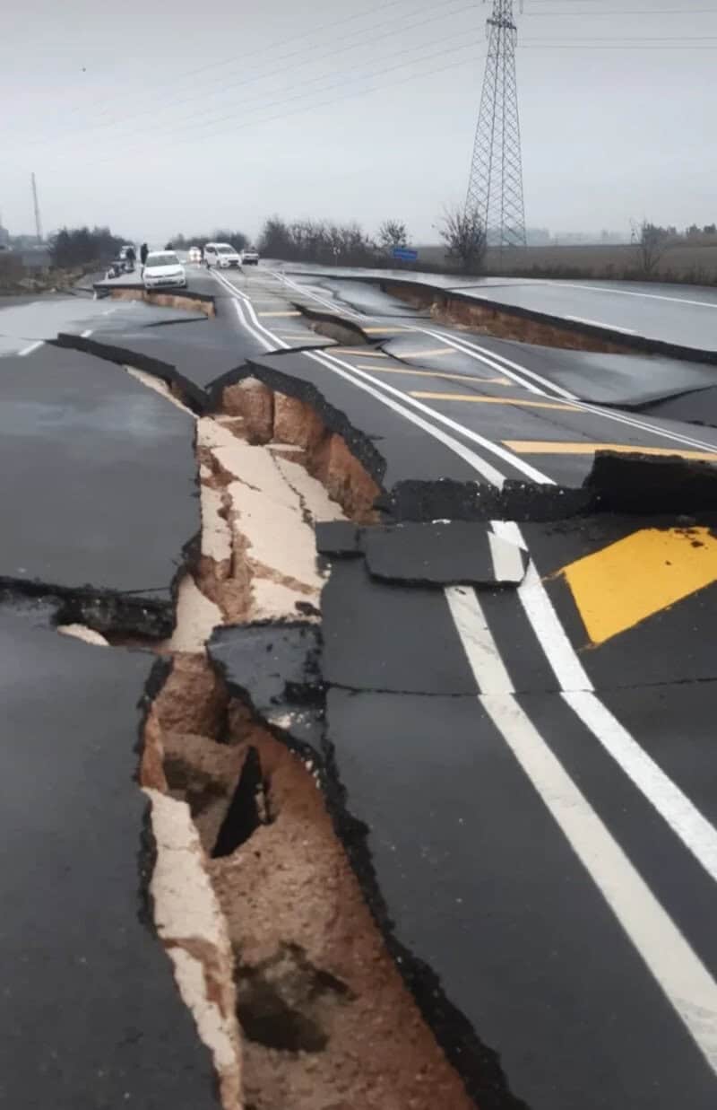 Damaged road with large cracks and collapsed sections, cars in the distance.