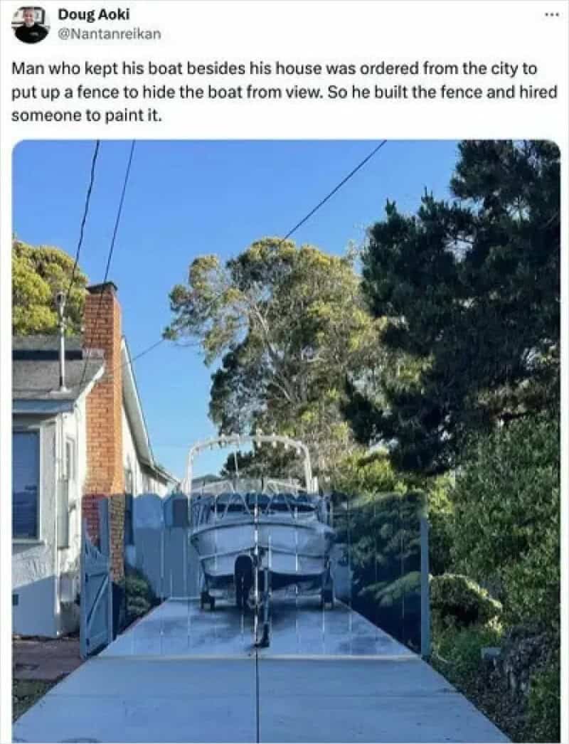 Boat built on a driveway, painted to hide it from view, with a fence in front.
