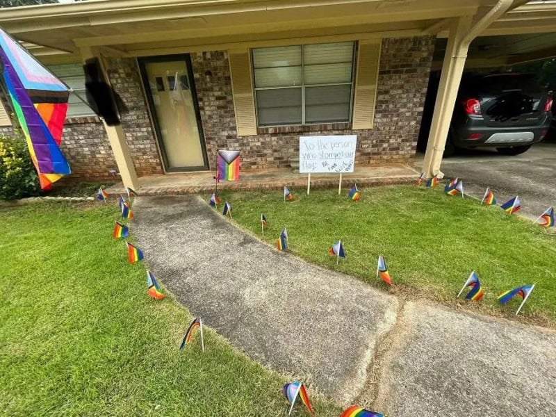 Colorful pride flags surround a concrete path leading to a house with a sign.