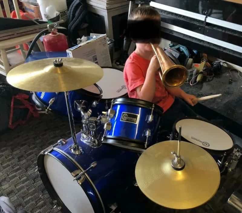 Child playing drum set and blowing into a trumpet in a cluttered room.
