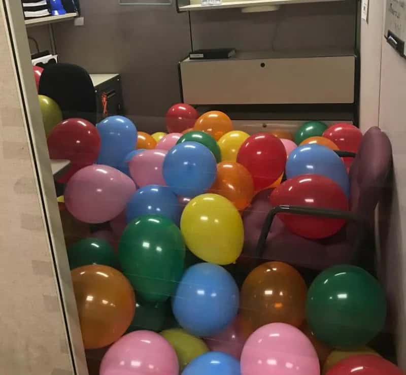 Room filled with colorful balloons, some on chairs and floor, with office furniture in background.