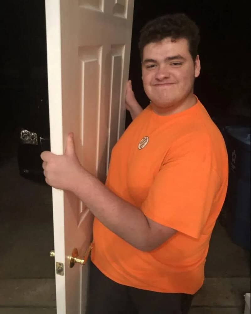 Young man in orange shirt smiling and holding a door open.