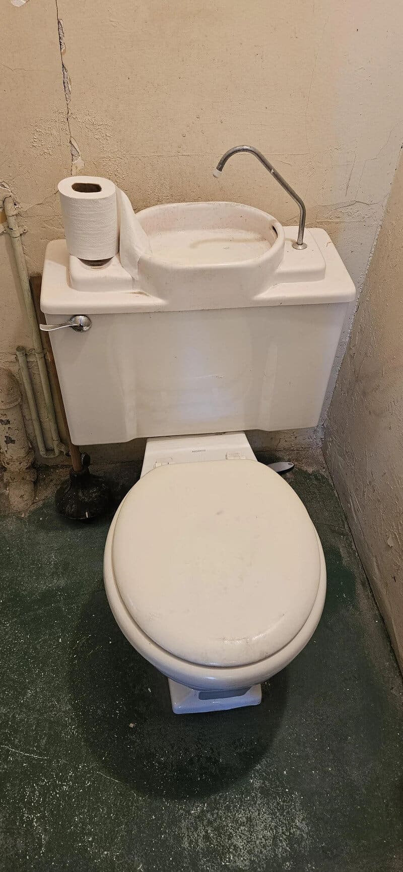 Toilet with sink on top, paper towel roll, and faucet in a small room.