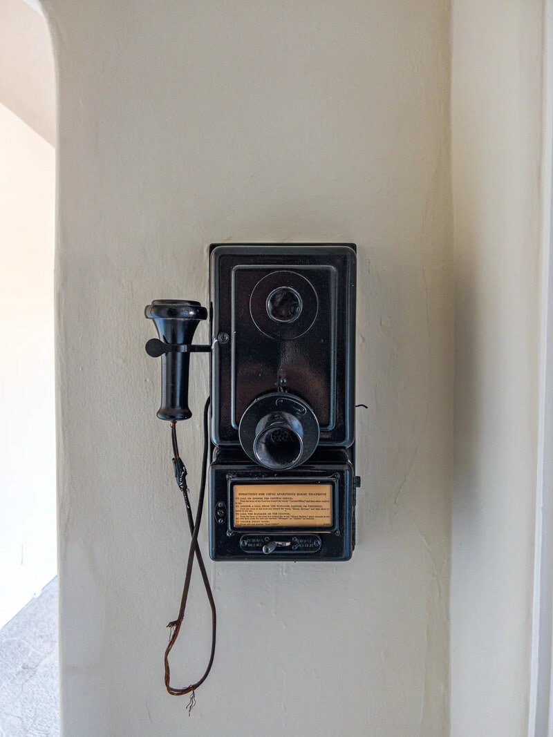Vintage black wall-mounted telephone with a rotary dial and cord on a beige wall.